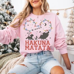 Lion King Hakuna Matata Christmas T-Shirt with Mickey Light Art 8 Female model in holiday bedroom wearing light pink crewneck fleece with glowing mickey light string outline tee graphic