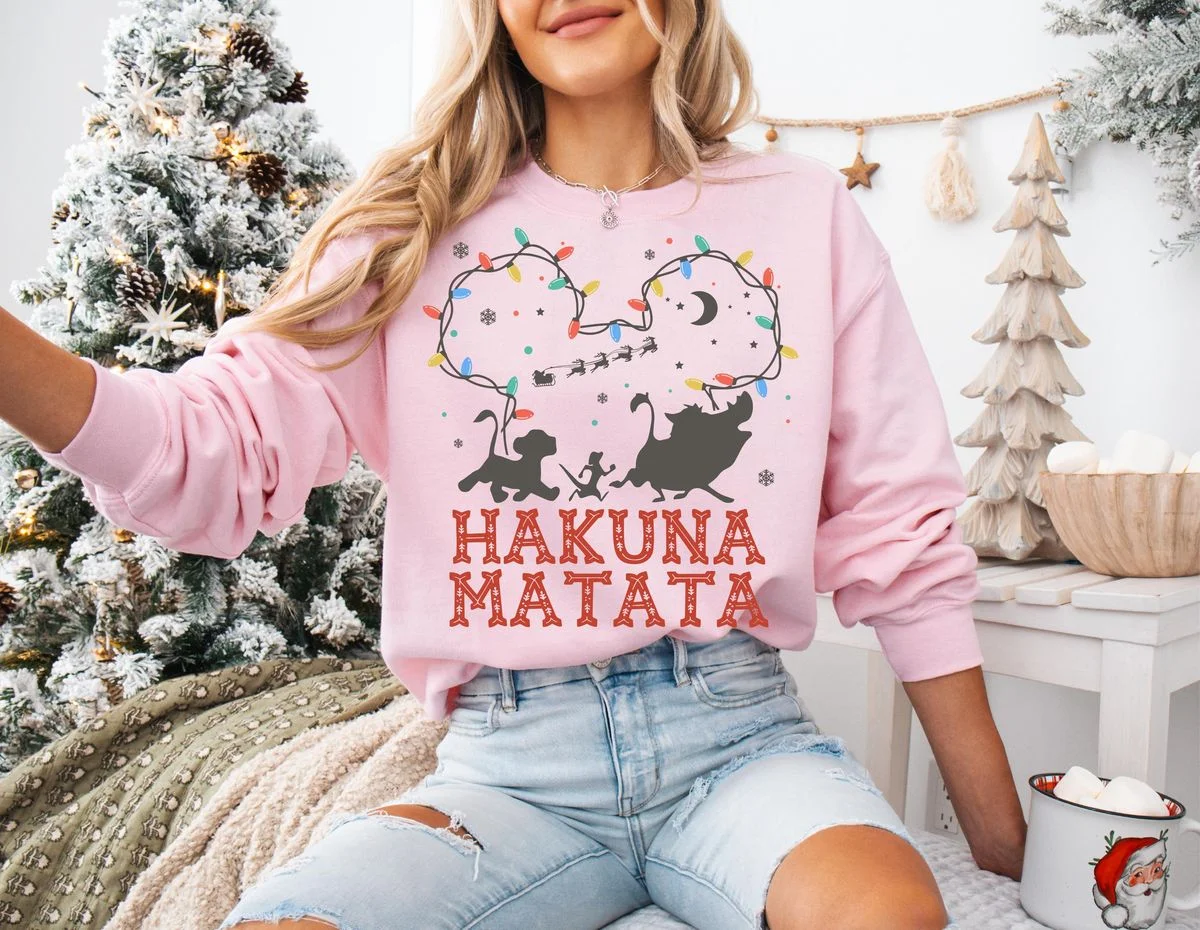 Lion King Hakuna Matata Christmas T-Shirt with Mickey Light Art 4 Female model in holiday bedroom wearing light pink crewneck fleece with glowing mickey light string outline tee graphic