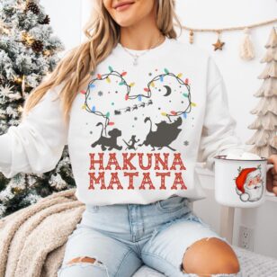 Lion King Hakuna Matata Christmas T-Shirt with Mickey Light Art 9 Woman sitting on white bedspread wearing white crewneck sweatshirt with Nordic Hakuna Matata holiday string light design