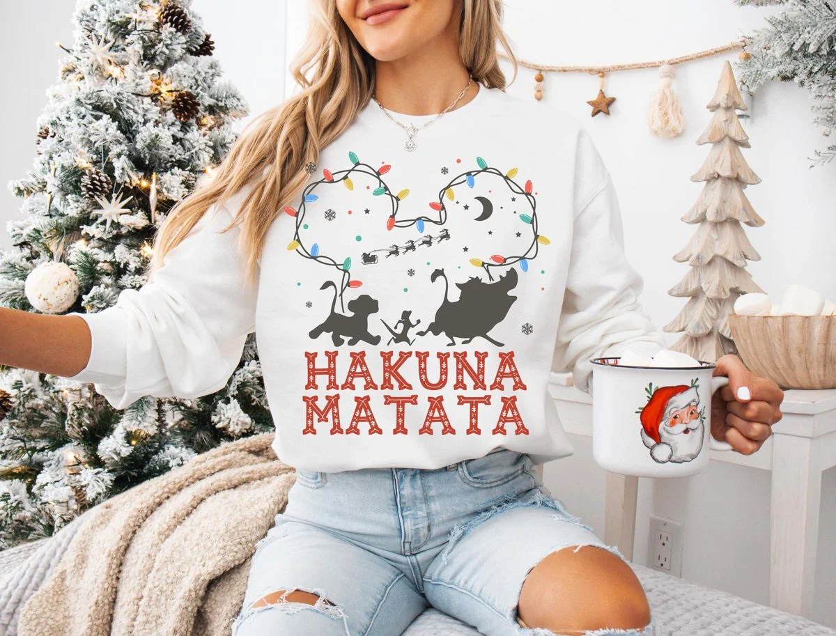 Lion King Hakuna Matata Christmas T-Shirt with Mickey Light Art 5 Woman sitting on white bedspread wearing white crewneck sweatshirt with Nordic Hakuna Matata holiday string light design