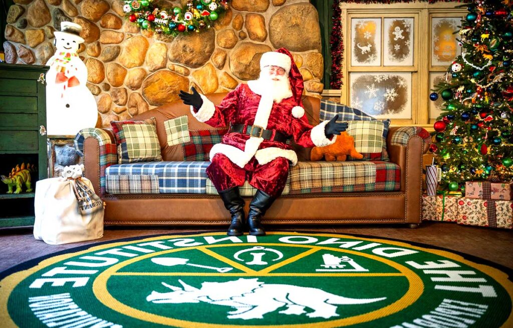 Christmas at Animal Kingdom Secrets Every Traveler Needs for 2027 17 Santa Claus sitting on a plaid couch over a green Dino Institute rug surrounded by dinosaur Christmas decorations at Animal Kingdom